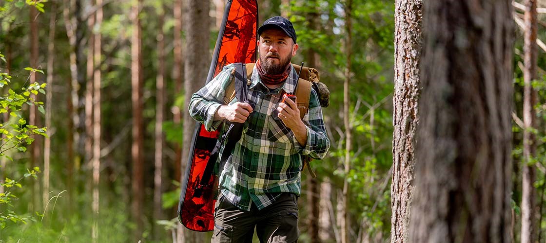 Mann går i skogen med våpenfutteral på ryggen og komradio i hånden.