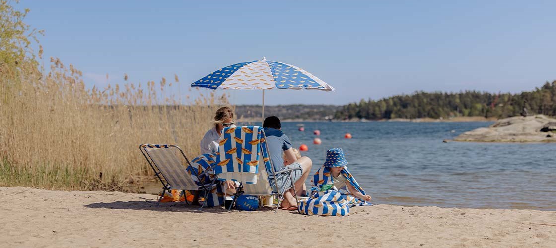 Bildet viser en familie på stranden. Mor sitter på et piknikk-pled og far sitter i en solstol. Barnen leker på stranden. 