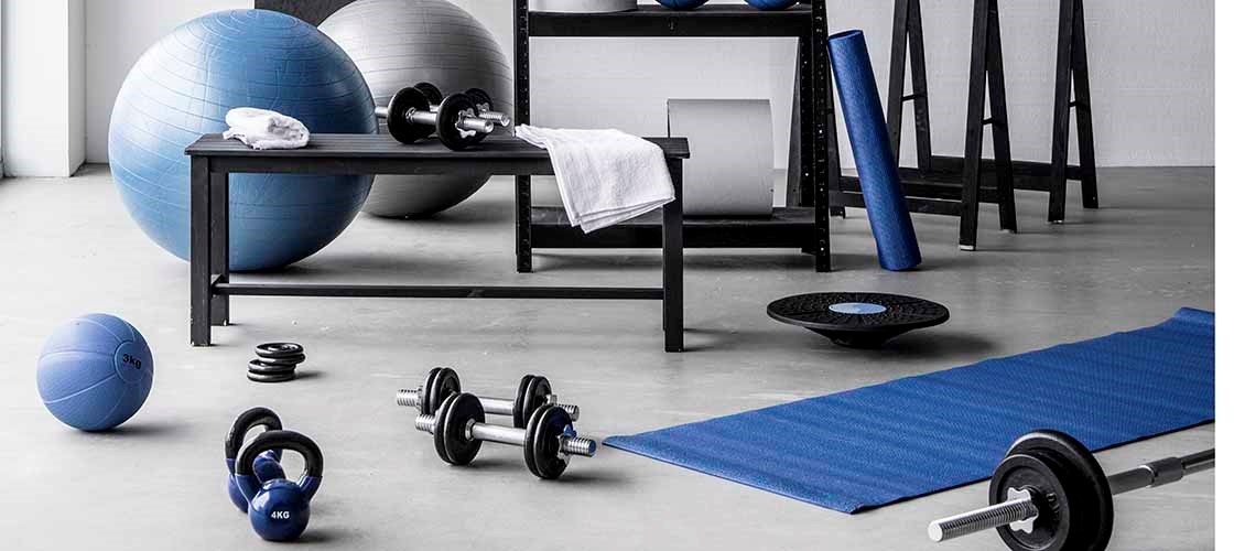 Bright and well-organized home gym featuring fitness equipment such as kettlebells, dumbbells, a barbell, exercise balls, and a yoga mat. The equipment is neatly stored on shelving and arranged across a modern workout space with white walls and a polished concrete floor. A handwritten “Squat Challenge” chart adds a motivational touch to the training environment.