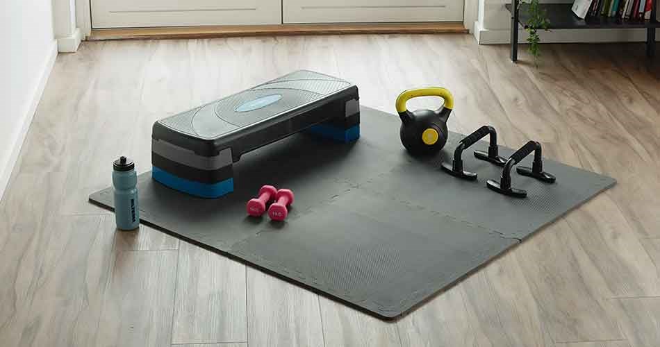 A workout mat with a step platform, kettlebell, dumbbells, push-up bars, and a water bottle is set up for home exercise.