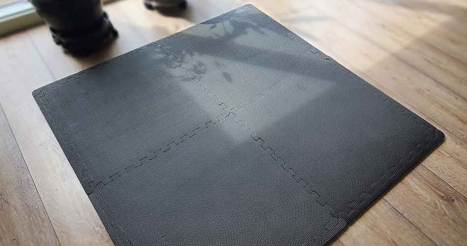 Four interlocking black foam training mats are placed on a wooden floor, forming a square workout surface.