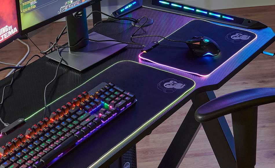 Gaming setup with RGB backlit keyboard and mouse on a black desk mat, surrounded by monitors and gaming equipment.