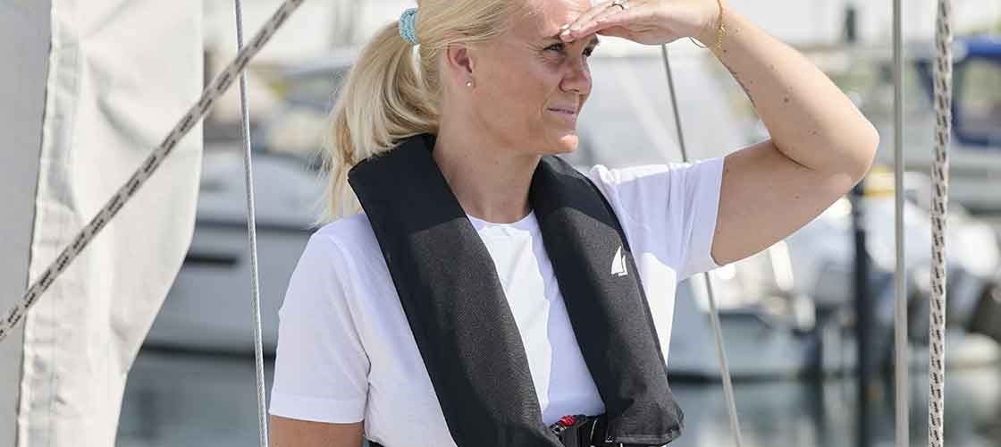Woman standing on the deck of a sailing ship wearing an inflatable life jacket. Used as inspiration for the guide: ‘How to check your inflatable life jacket - step by step’.