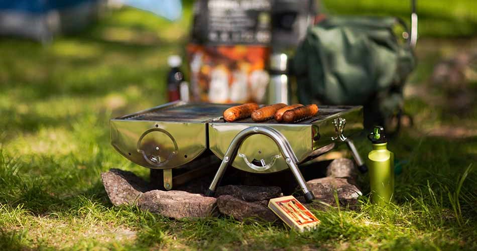 A portable grill is placed on the ground in a green area and is used to grill sausages.
The grill is set up on small legs on top of rocks and is surrounded by grass and outdoor equipment.
In the background, a bag, a bottle and packaging are visible, indicating an excursion or camping trip.