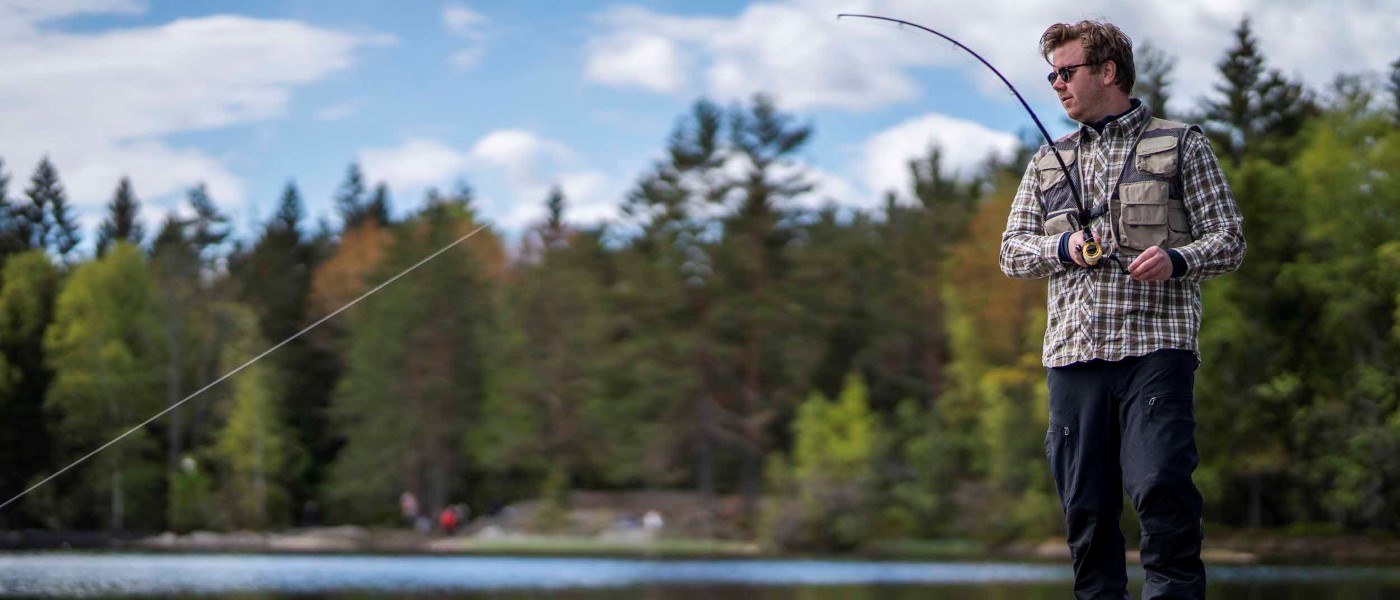 En person står ved vannkanten med en fiskestang og fisker i et naturområde. Personen har på seg rutete skjorte, vest og solbriller, mens fiskestangen er bøyd fremover. Trær og grønn vegetasjon er synlige i bakgrunnen under en delvis skyet himmel.