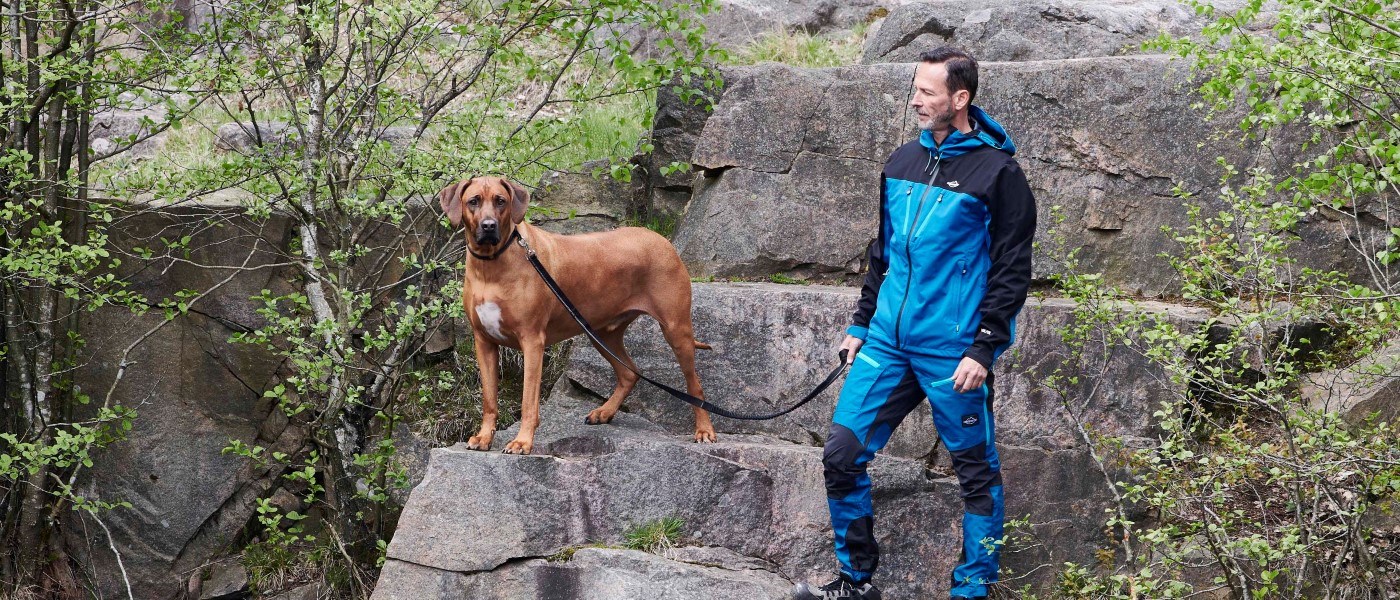 En person i blått friluftstøy står på steiner i et grønt naturområde og holder en hund i bånd. Hunden står på en steinhylle og ser fremover, mens båndet går mellom hunden og personen. Store steinblokker, busker og trær omgir stedet.