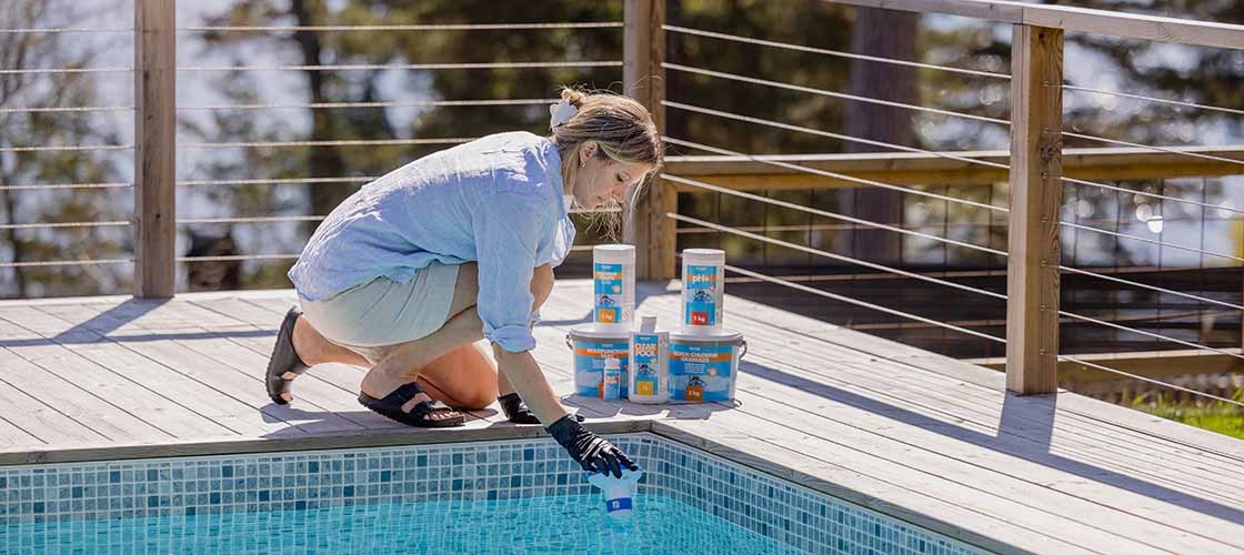 A person wearing a protective glove tests the pool water at the edge of an outdoor pool. Several containers of pool chemicals stand on the wooden deck next to the pool.