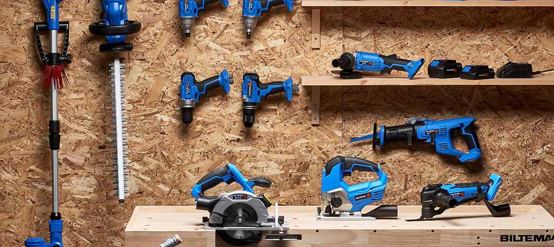 A plywood-clad wall where a range of blue and black power tools from Biltema are hung and displayed, including drills, circular saws and angle grinders. Additional tools and batteries are displayed on a shelf.